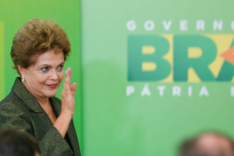Dilma aparece acenando, do peito para cima. Ela tem cabelos de cor laranja, veste paletó verde escuro e usa brincos.