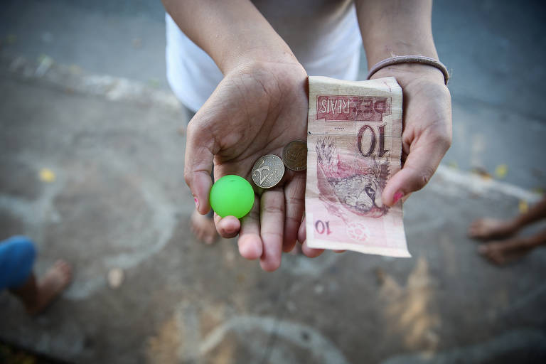 Criança segura na mão uma cédula de dinheiro, algumas moedas e uma bola pequena 