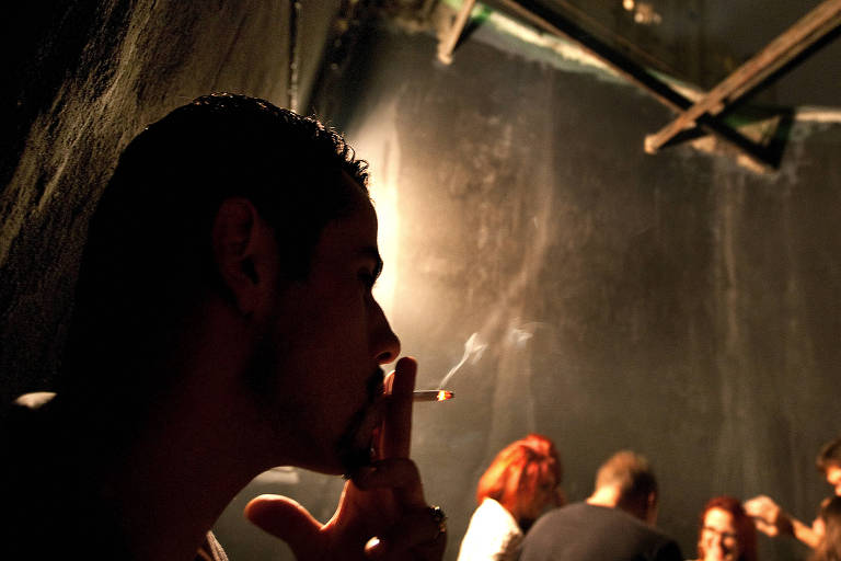 Silhueta de homem fumando cigarro em primeiro plano, com pessoas conversando ao fundo, em local que parece fumódromo de uma casa noturna