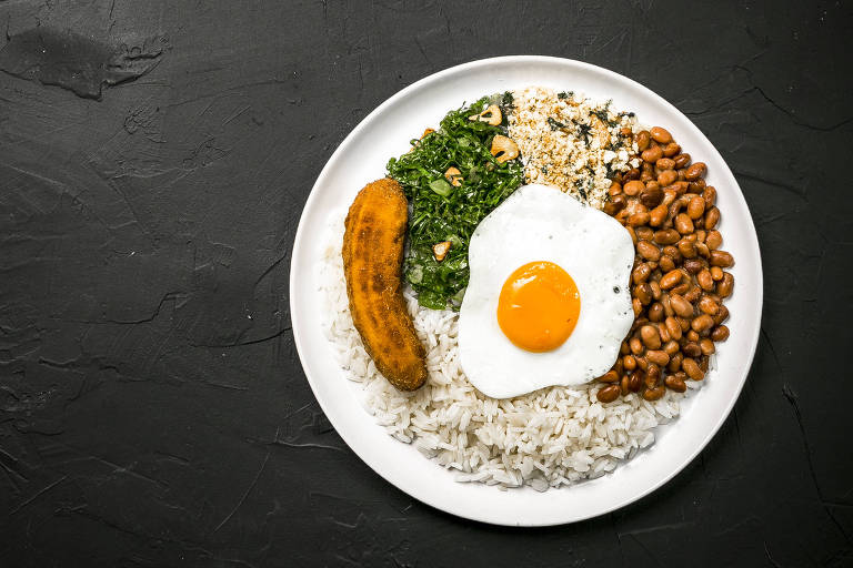 Um prato redondo com arroz, feijão, um pedaço de carne, folhas verdes e um ovo frito no centro. O fundo é escuro, destacando o prato e os alimentos.