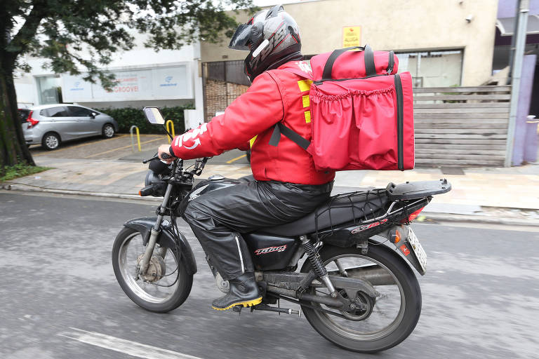 Homem de jaqueta vermelha e calça preta andando em uma moto com uma mochila de entrega vermelha e capacete preto