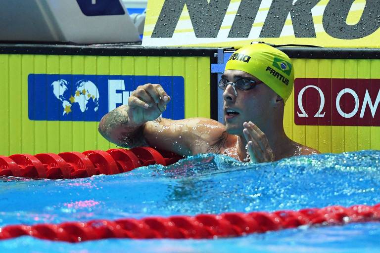 Nadador dentro da piscina, submerso até a altura do peito, reage ao saber que conquistou o segundo lugar da prova. Utiliza touca amarela e óculos de natação pretos. 