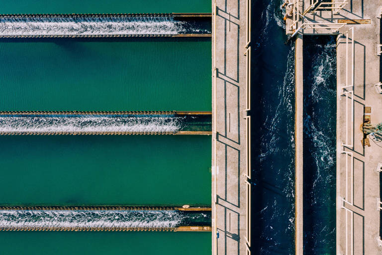 A imagem captura uma vista aérea de uma barragem, mostrando a água em movimento criando padrões de espuma branca enquanto flui através das comportas. A composição simétrica divide a fotografia, com a estrutura da barragem formando uma linha divisória.
