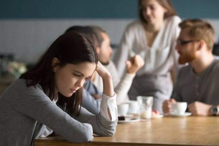em primeiro plano, mulher jovem apoia a cabeça com os cotovelos e está sentada à mesa com expressão triste. Ao fundo, jovens conversam e exibem expressões de animação.
