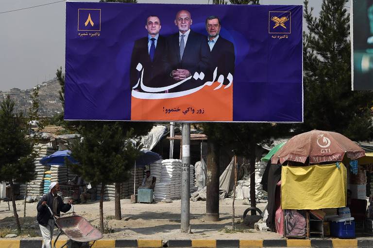 Um morador local empurra um carrinho de mão por um outdoor com um cartaz do candidato presidencial afegão e do atual presidente do Afeganistão, Ashraf Ghani.