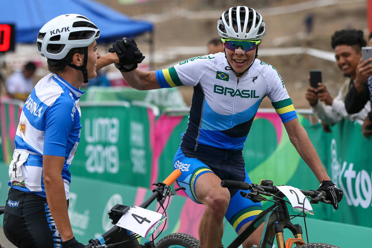 Jaqueline Mourão sorri em cima da bicicleta e receber um cumprimento de outra atleta ao cruzar a linha de chegada