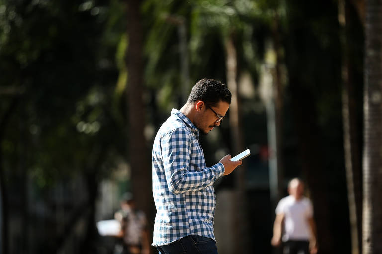 Um homem está de perfil, olhando para o celular enquanto caminha em um ambiente urbano. Ele usa uma camisa xadrez azul e branca e óculos. Ao fundo, há pessoas caminhando e árvores, sugerindo um dia ensolarado.