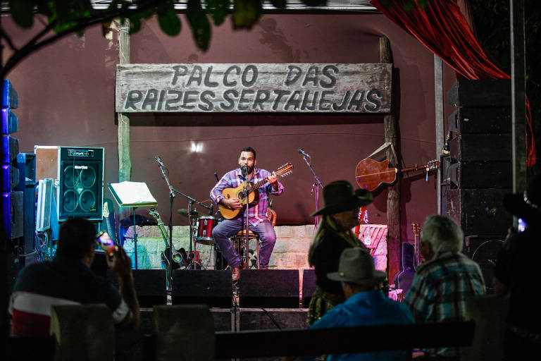 Homem com violão em palco se apresenta para plateia vazia. Atrás dele, uma placa diz Palco das Raízes Sertanejas