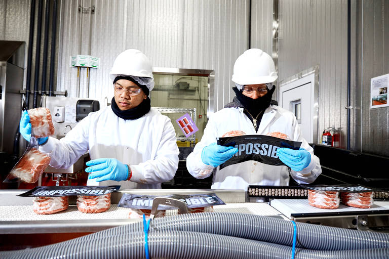 Operários manipulam embalagens com hambúrgueres vermelhos, sobre esteira com diversas outras embalagens