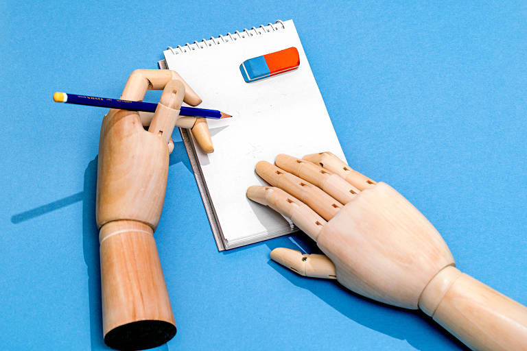 Foto mostra um fundo azul com um bloco de anotações de folhas brancas com uma borracha azul e vermelha apoiada sobre ele. Um mão articulada de madeira segura a caderneta e outra mãos articulada de madeira segura um lápis azul posicionado para escrever.