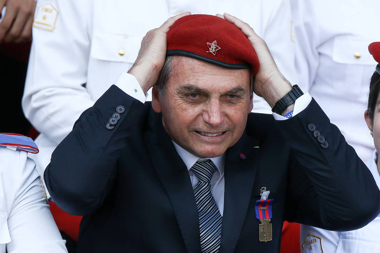 Bolsonaro veste uma boina vermelha do exército com um meio sorriso e segura a peça com as mãos sobre a cabeça