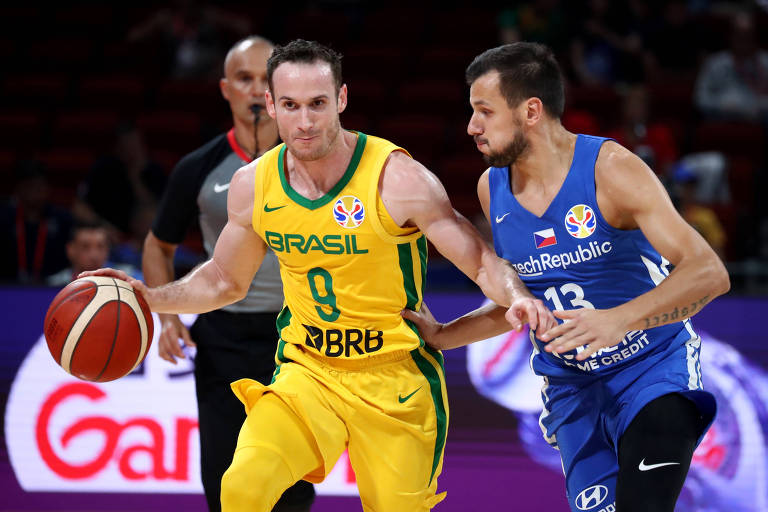 à esquerda, jogador de basquete domina a bola com a mão direita e afasta o adversário com o braço esquerdo. Marcelinho Huertas é da seleção brasileira e traja uniforme amarelo. À direita, jogador da República Tcheca, com traje azul, olha para a bola de basquete e marca o adversário