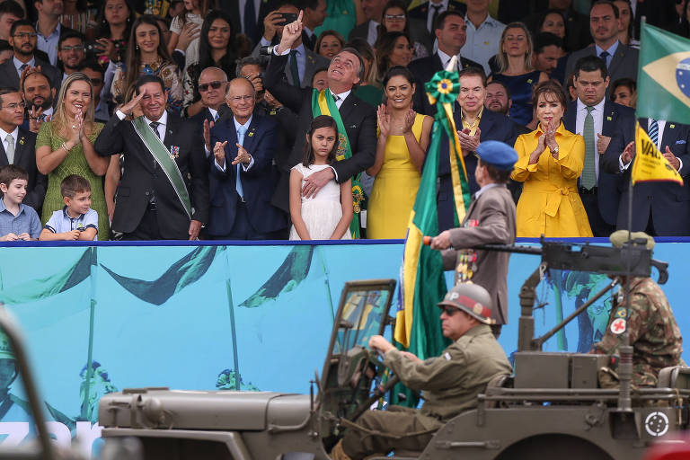 Presidente Bolsonaro aponta para o alto, segurando a filha à sua frente, e vice Mourão bate continência para carro militar que desfila 
