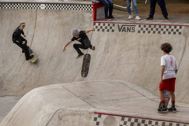 Três dolescentes andam de skate na pista Vans Skatepark, mais desenhada e elaborada para o esportista fazer manobras, com obstáculos e também "bowls", uma espécie de parede côncava, no parque Cândido Portinari, vizinho do Villa-Lobos. O jovem na parte central da imagem realiza uma manobra no ar