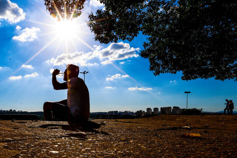 A imagem mostra a silhueta de uma pessoa sentada no chão， segurando um objeto， possivelmente uma câmera ou celular， enquanto observa o céu. O sol brilha intensamente atrás dela， criando um efeito de raios solares. O céu é azul com nuvens brancas e há árvores ao redor. Ao fundo， é possível ver um horizonte urbano com edifícios e outras pessoas caminhando.