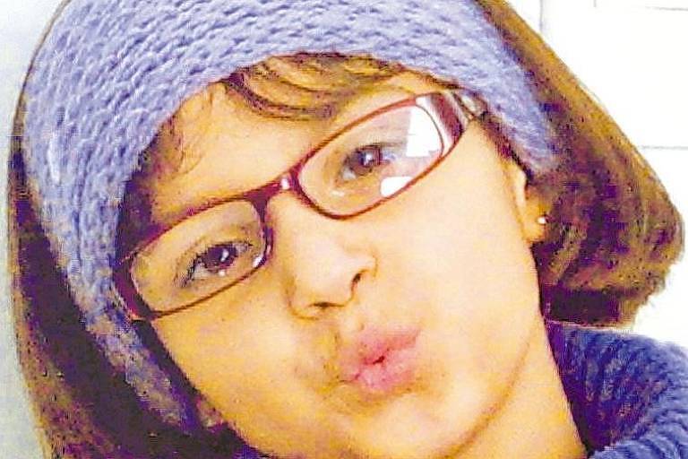 Foto mostra menina de cabelos na altura do queixo, franjinha, que usa óculos e roupa de frio, inclusive uma faixa de lã na cabeça, em tons de roxo e lilás; ela faz um beijinho com a boca para a câmera