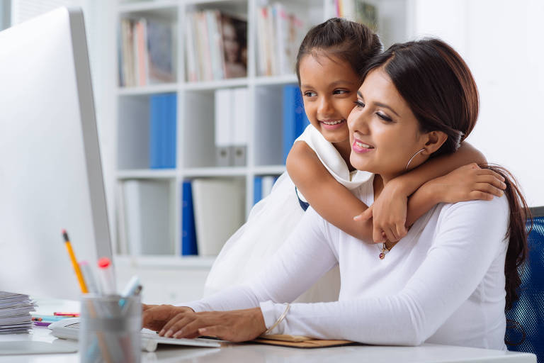 Mulher é abraçada por criança, enquanto está em frente ao computador