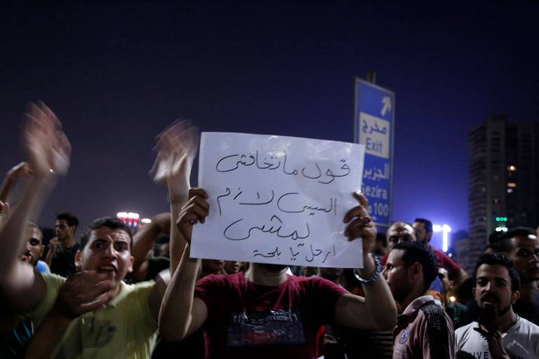 Ativistas seguram cartazes em árabe e gesticulam com as mãos para cima, na noite da praça Tahir, no Cairo. 