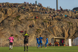 Fans watch from the top of a cliff that serves as a grandstand as players on the team G-44 (blue) celebrate a goal against IT-79 (pink) in Sisimiut, Greenland, Aug. 8, 2019. (Kieran Dodds/The New York Times)