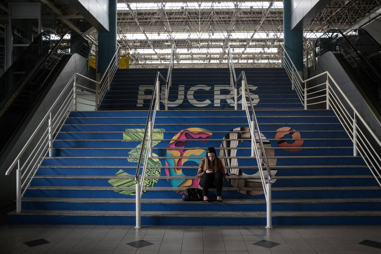 Aluna sentada em escadaria da Pontifícia Universidade Católica do Rio Grande do Sul. Nos degraus azuis estão pintados os dizeres "PUCRS" em branco e "360°" em várias cores