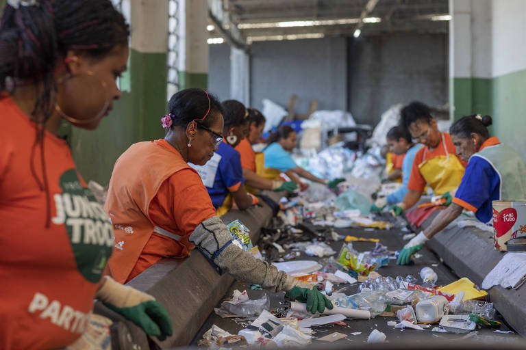 Foto mostra trabalhadores catando objetos de uma esteira cheia de lixo, dentro de um galpão.