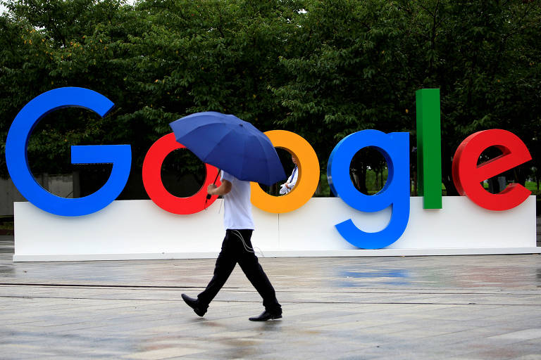 Homem com a cabeça encoberta por um guarda-chuva azul marinho caminha em frente a grandes letras coloridas que formam a palavra Google. Ele está passando justamente entre as duas letras o, o que cria um efeito visual interssante