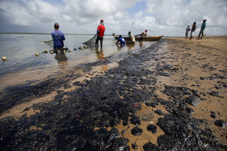 Manchas de óleo na areia com pescadores segurando redes ao fundo