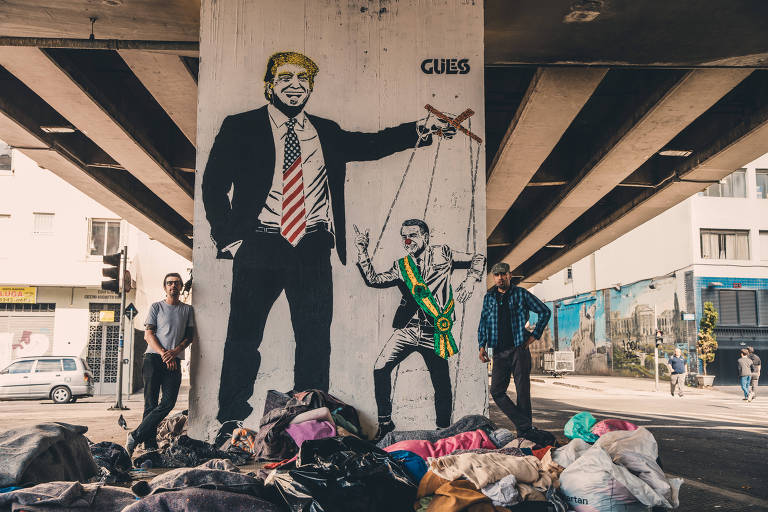 Grafiteiro Guiless (camisa xadrez) e seu parceiro Iano Ahmed (óculos preto) fazem charge política na pilastra do Minhocão, em que Trump manipula Bolsonaro como um fantoche ou marionete