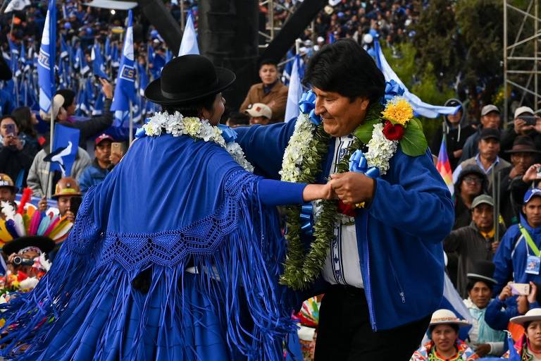 Usando colar indígena, presidente dança segurando as mãos de uma mulher com roupa azul. É observado por apoiadores