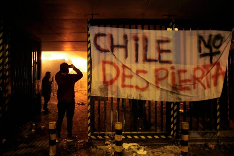 Estação do metrô incendiada pelos manifestantes, no sábado (19); ao lado um cartaz que diz: 'Chile não acorda'.
