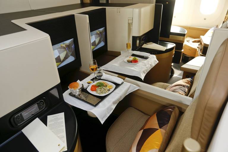 A imagem mostra uma cabine de primeira classe de uma companhia aérea, com assentos espaçosos que se convertem em camas, equipados com telas de entretenimento pessoais. Uma refeição gourmet está sendo servida em uma bandeja, acompanhada de uma taça de vinho.