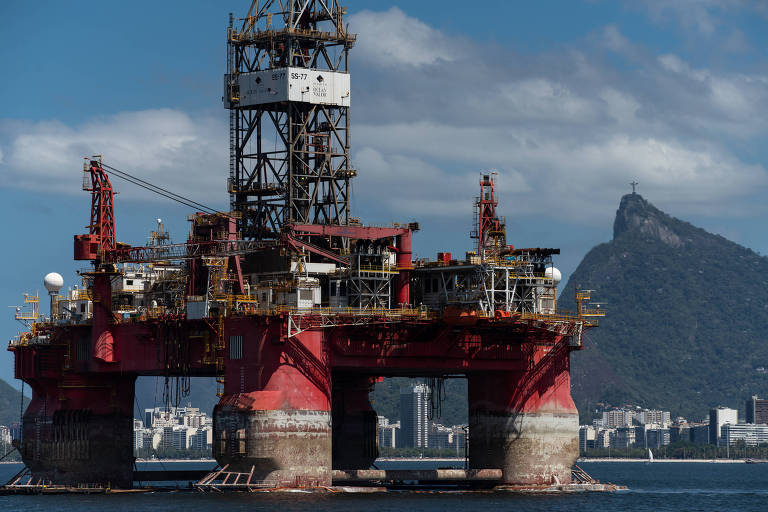 A imagem mostra uma plataforma de perfuração de petróleo em águas do Rio de Janeiro, com um céu azul e algumas nuvens. Ao fundo, é possível ver uma montanha com o Cristo Redentor no topo e uma área urbana com prédios à beira-mar.
