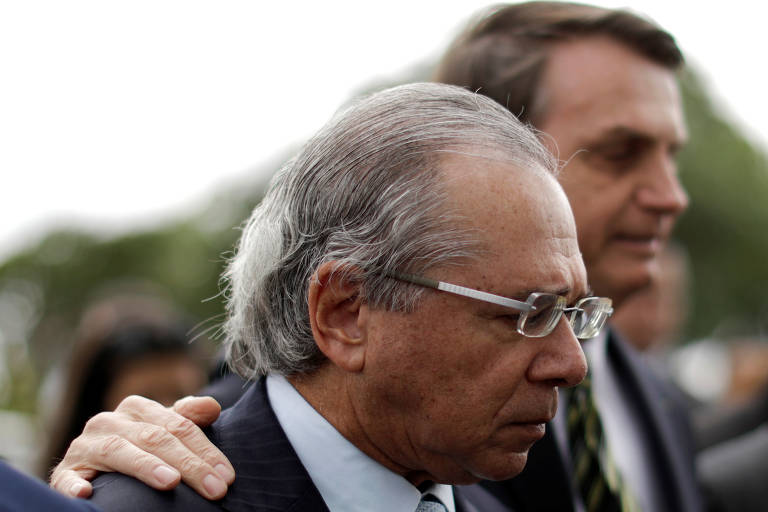 Paulo Guedes olha para baixo, franzindo a testa. Ao seu lado e ao fundo, Jair Bolsonaro, com rosto desfocado, repousa a mão no ombro do ministro.