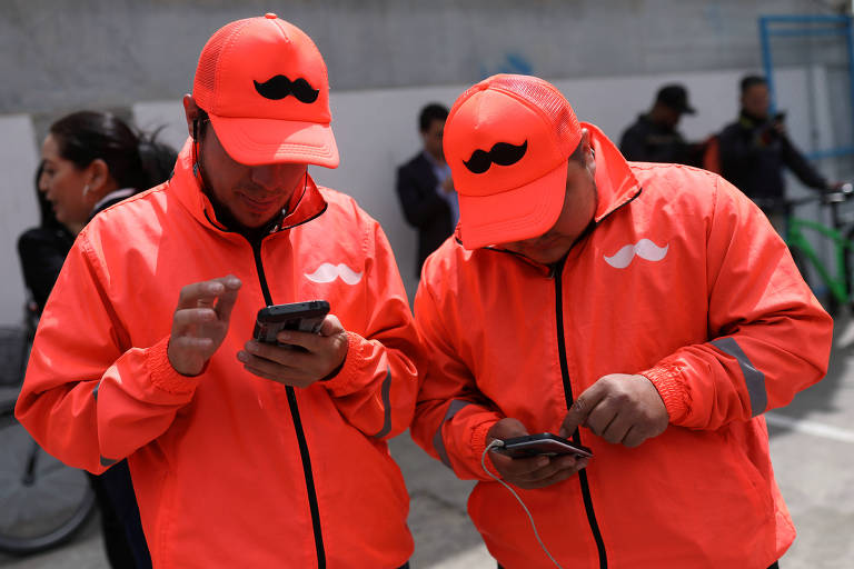 Dois homens com jaquetas e bonés da empresa de entregas Rappi olham seus celulares na rua, em Bogotá, na Colômbia