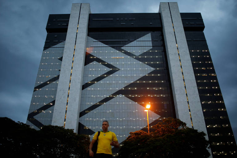 Homem vestido de amarelo caminha em frente a um prédio, cuja fachada é tomada pelo símbolo do Banco do Brasil