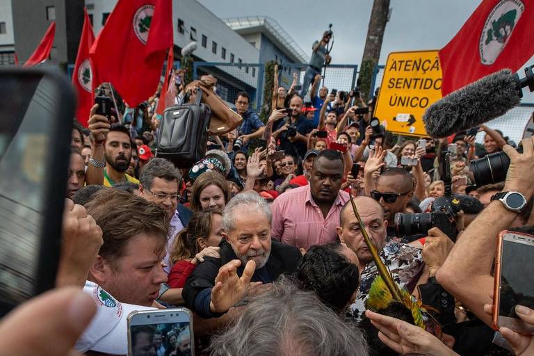 Na foto Lula ao centro, rodeado por apoiadores, seguranças, jornalistas e fotógrafos. Entre as dezenas de apoiadores, bandeiras do Movimento sem Terra (MST). Ao fundo, Superintendência da Polícia Federal.