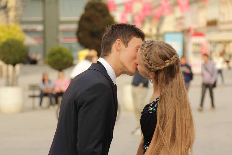 Um homem e uma mulher se beijam de uma forma discreta, em uma praça, durante o dia. Não é possível ver o rosto dos dois, apenas os cabelos e perfis. A mulher tem o cabelo loiro e o homem tem o cabelo curto e castanho escuro