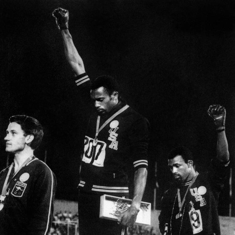 Os atletas negros Tommie Smith (centro) e John Carlos (dir.), medalhas de ouro e de bronze nos 200 m, fazem a saudação com o punho cerrado do grupo Panteras Negras, como protesto a conflitos raciais nos EUA