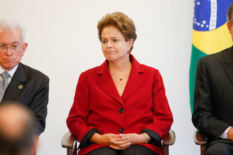Dilma aparece sentada, da cintura para cima. Ela tem cabelos de cor alaranjada, entrelaça os dedos sobre as pernas e olha para o lado, vestindo paletó vermelho. À sua esquerda e direita, encontram-se outros membros do governo sentados. Atrás do que está à sua esquerda, aparece parte da bandeira do Brasil.