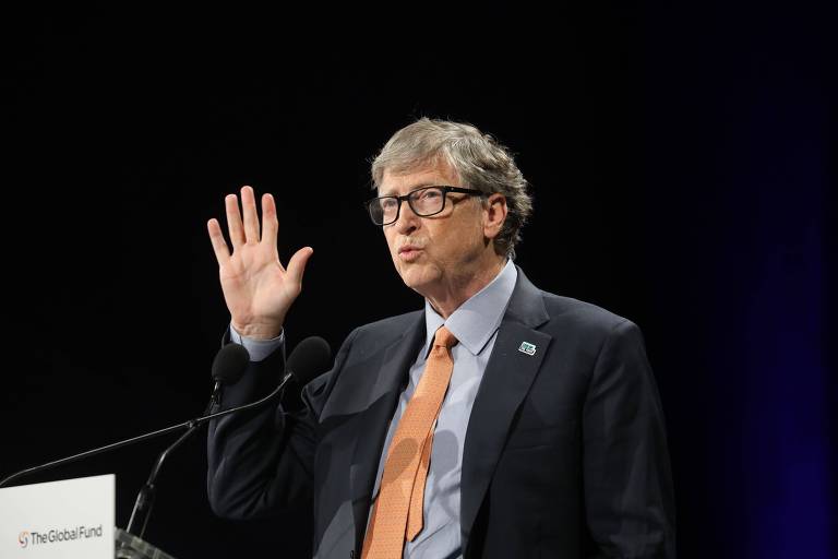 Na foto, o fundador da Microsoft, Bill Gates, aparece em um palanque falando em uma palestra, com uma das mãos erguida no ar.