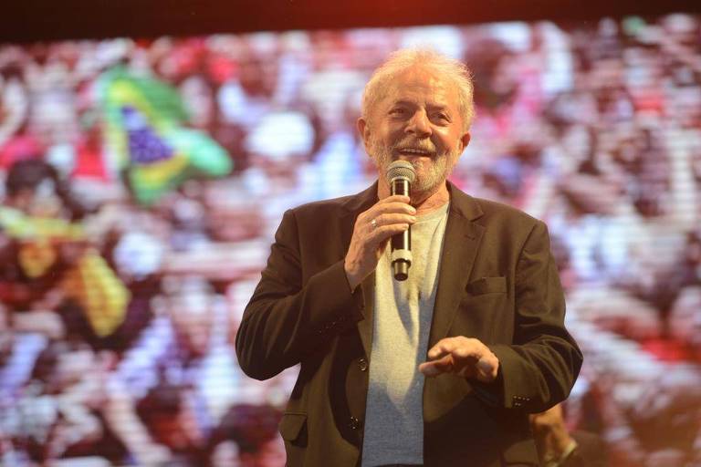 Lula segura o microfone e sorri.Ele veste um blazer preto e uma camisa cinza. Atrás, o telão mostra a plateia, majoritariamente vestida de vermelho