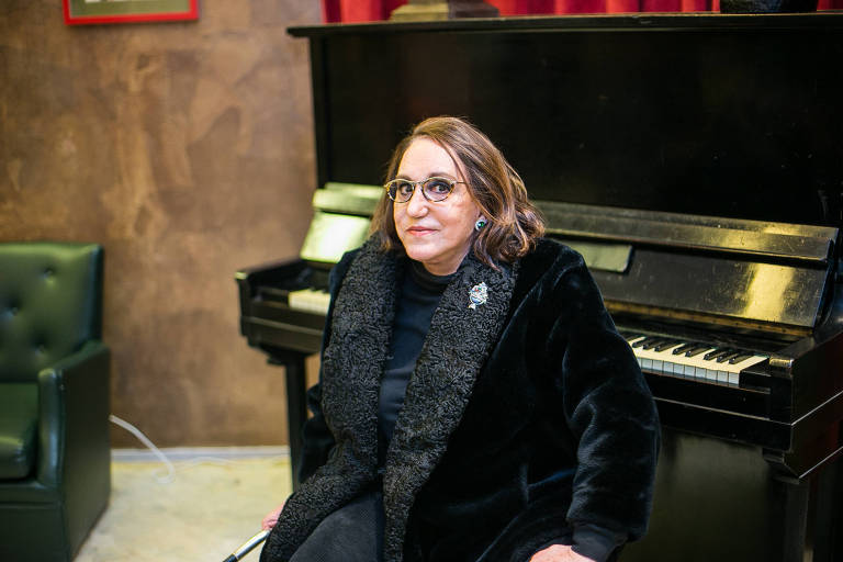 A autora Leilah Assumpção, vestindo um casaco preto, sentada em frente a um piano