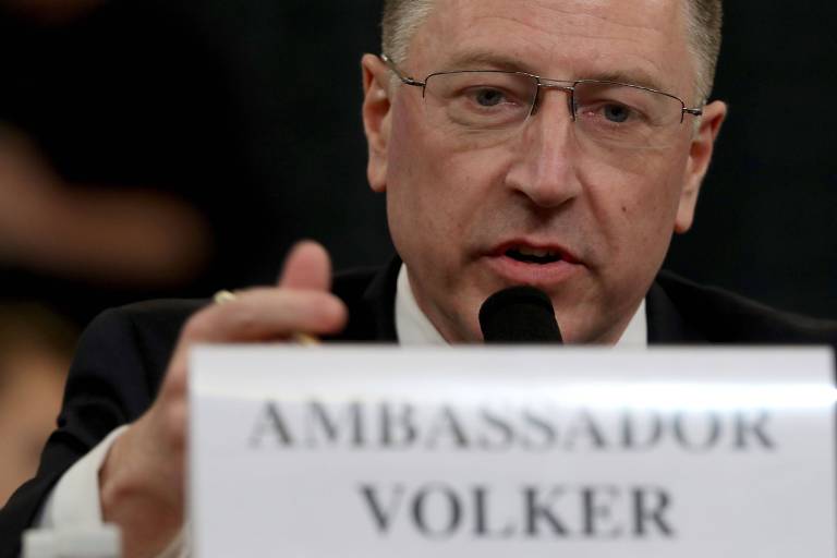 Kurt Volker gesticula enquanto fala em um microfone; a sua frente, há uma placa branca onde se lê "ambassador Volker" (embaixador Volker)