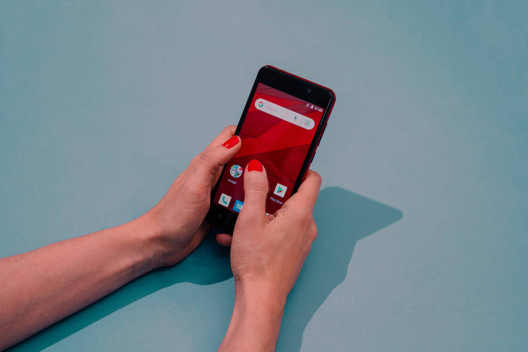 Mãos com as unhas pintadas de vermelho estão segurando um celular com a tela desbloqueada