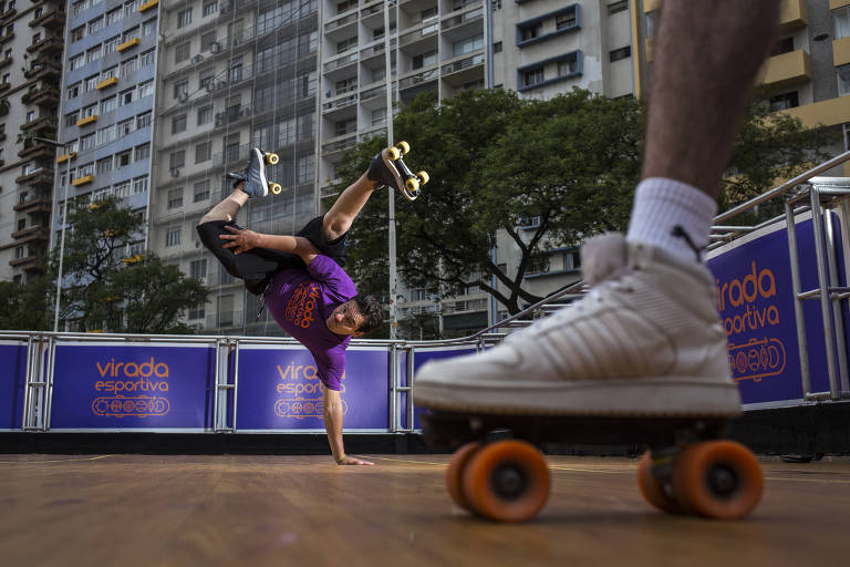 foto mostra em primeiro plano o pé de uma pessoa sobre um skate. em segundo plano, do lado esquerdo, uma pessoa pratica manobra de skate com as pernas para cima, apoiando o braço esquerdo no chão 