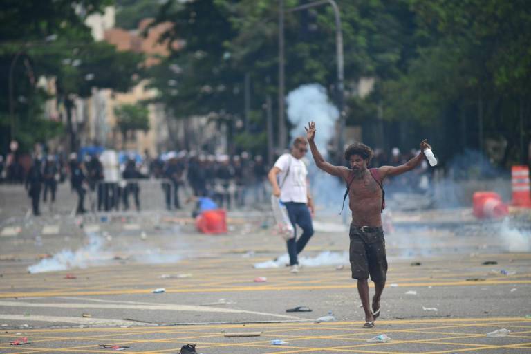Num ambiente de desordem, homem sem camisa e com braços erguidos caminha na rua após confronto com a polícia