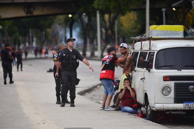 Policial atende torcedores acuados próximo a um veiculo na beira da calçada