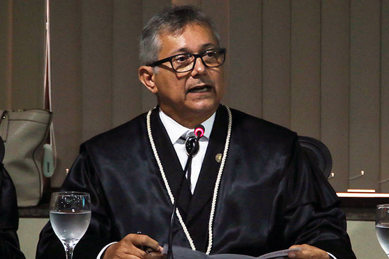 O procurador do Ministério Público do Pará Ricardo Albuquerque da Silva veste óculos e toga. Ele fala ao microfone enquanto está sengado à mesa. Há um copo de água ao seu lado direito. Ele segura um papel e olha para o lado direito da imagem. 