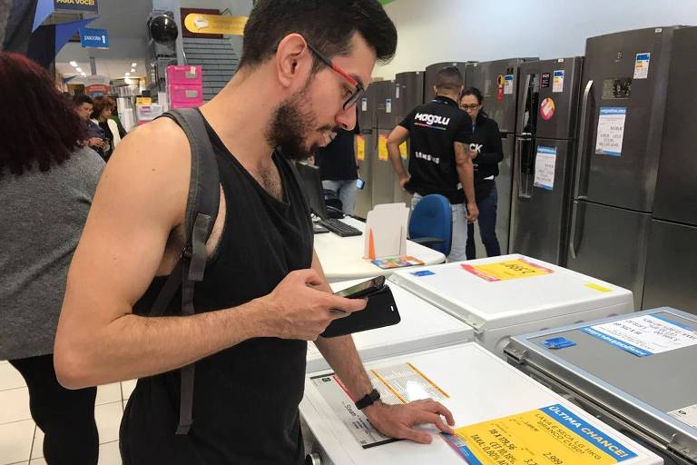 Um homem está parado na frente de uma máquina de lavar e segura um celular em uma das mãos, comparando o preço do produto na internet.