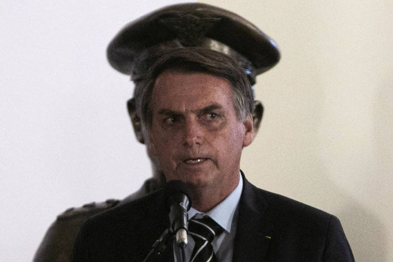 Jair Bolsonaro fala ao microfone; atrás dele, uma estátua de um busto militar. 